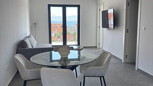 Uma sala de jantar moderna com mesa de vidro e quatro cadeiras, um sofá e uma TV na parede.