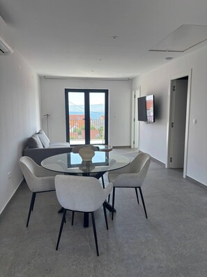 Ein moderner Essbereich mit einem Glastisch, vier Stühlen, einer Couch und einem an der Wand befestigten Fernseher.