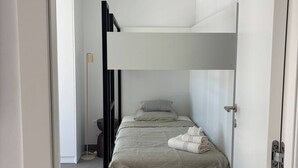 Uma cama de beliche com cabeceira branca, uma mesinha de cabeceira e um abajur.