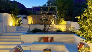 Uma área de estar moderna ao ar livre, com um banco embutido, uma mesa e cadeiras, cercada por um muro de pedra e uma árvore.