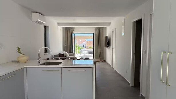 A modern kitchen with a central island, a built-in oven, and a sink. There is a view of a balcony with a railing and a building in the background.
