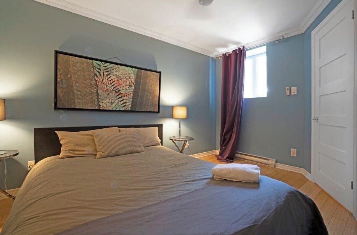 Une chambre à coucher avec un lit, une table de chevet, une lampe, une fenêtre avec des rideaux et une porte.