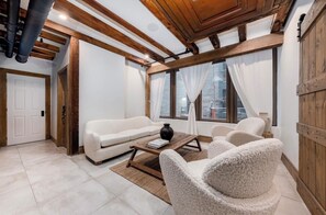 Een woonkamer met een witte bank, een bijzettafeltje en twee fauteuils. Het plafond is van hout met zichtbare balken en er is een groot raam met gordijnen.