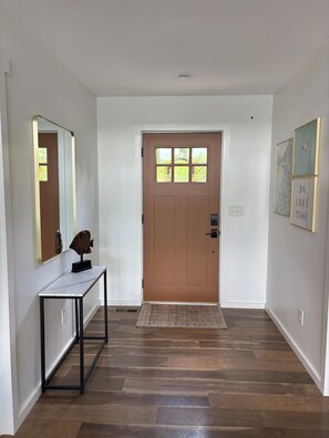 A hallway with a wooden floor, a brown door, a white wall with a mirror, and a framed picture. - Fresh and Modern Lake House Steps to the Water! (Dry Ridge)