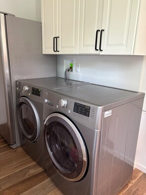 A compact, stainless steel front-loading washer and dryer unit with a digital display and control panel, installed in a kitchen with white cabinets and a wooden floor.