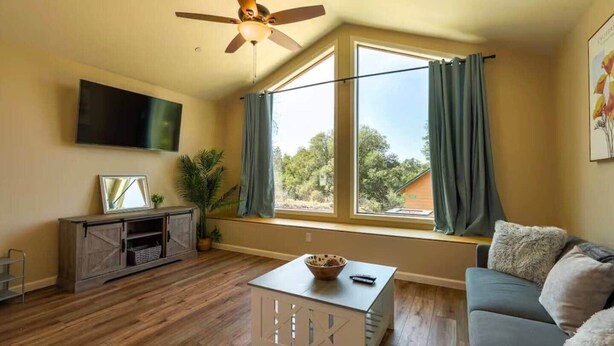 A living room with a ceiling fan, a TV mounted on the wall, a sofa, a coffee table, and a window with curtains. - Ponerosa Nest-Quails Nest 1hr Drive to Yosemite (Mariposa County)