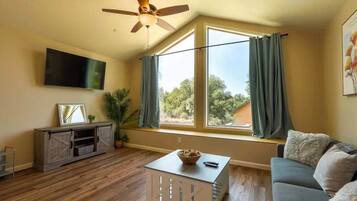 Un salon avec un ventilateur de plafond, un téléviseur fixé au mur, un canapé, une table basse et une fenêtre avec des rideaux.