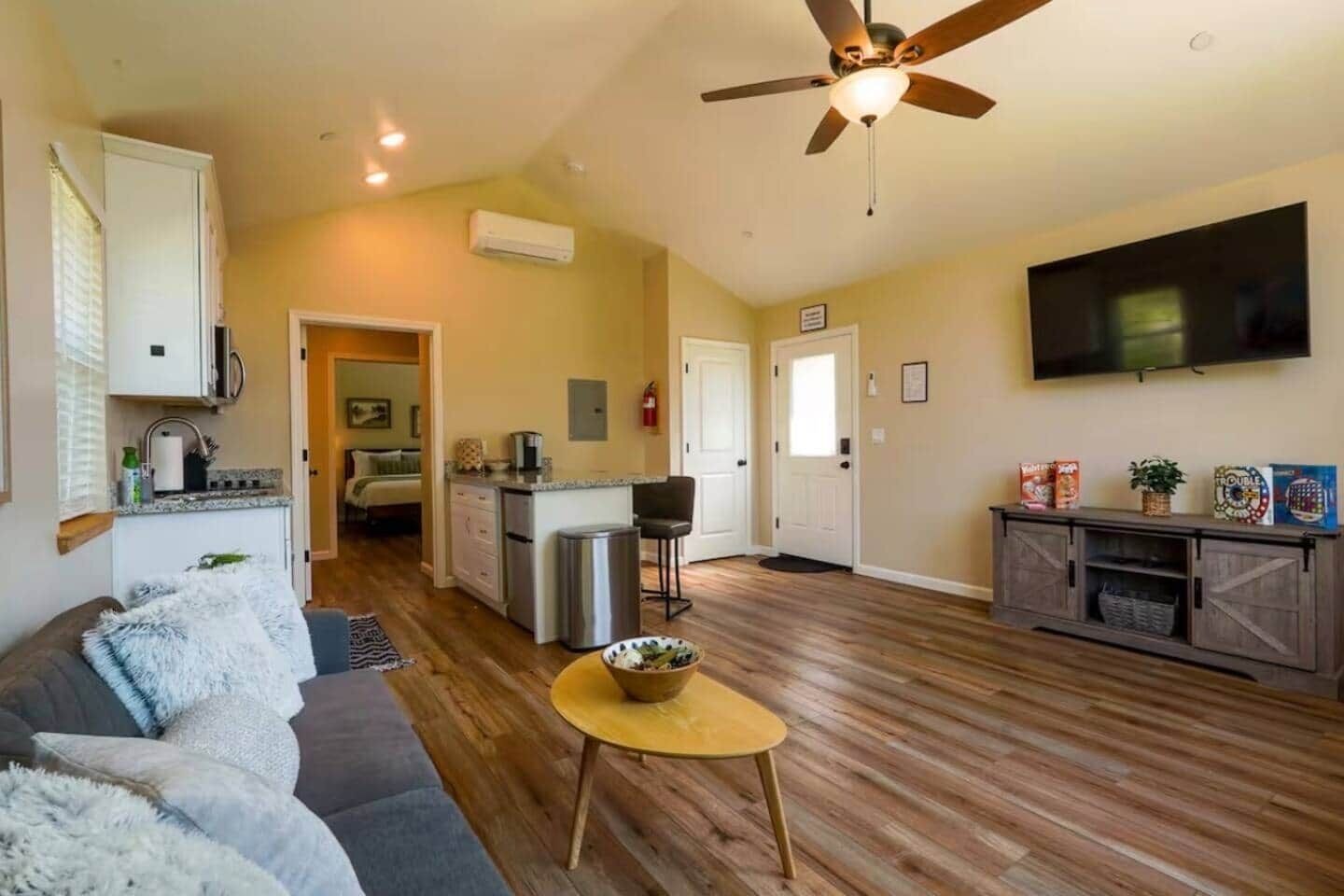 A living room with a sofa, a coffee table, a TV, a ceiling fan, and a kitchen area with a fridge, microwave, and sink.