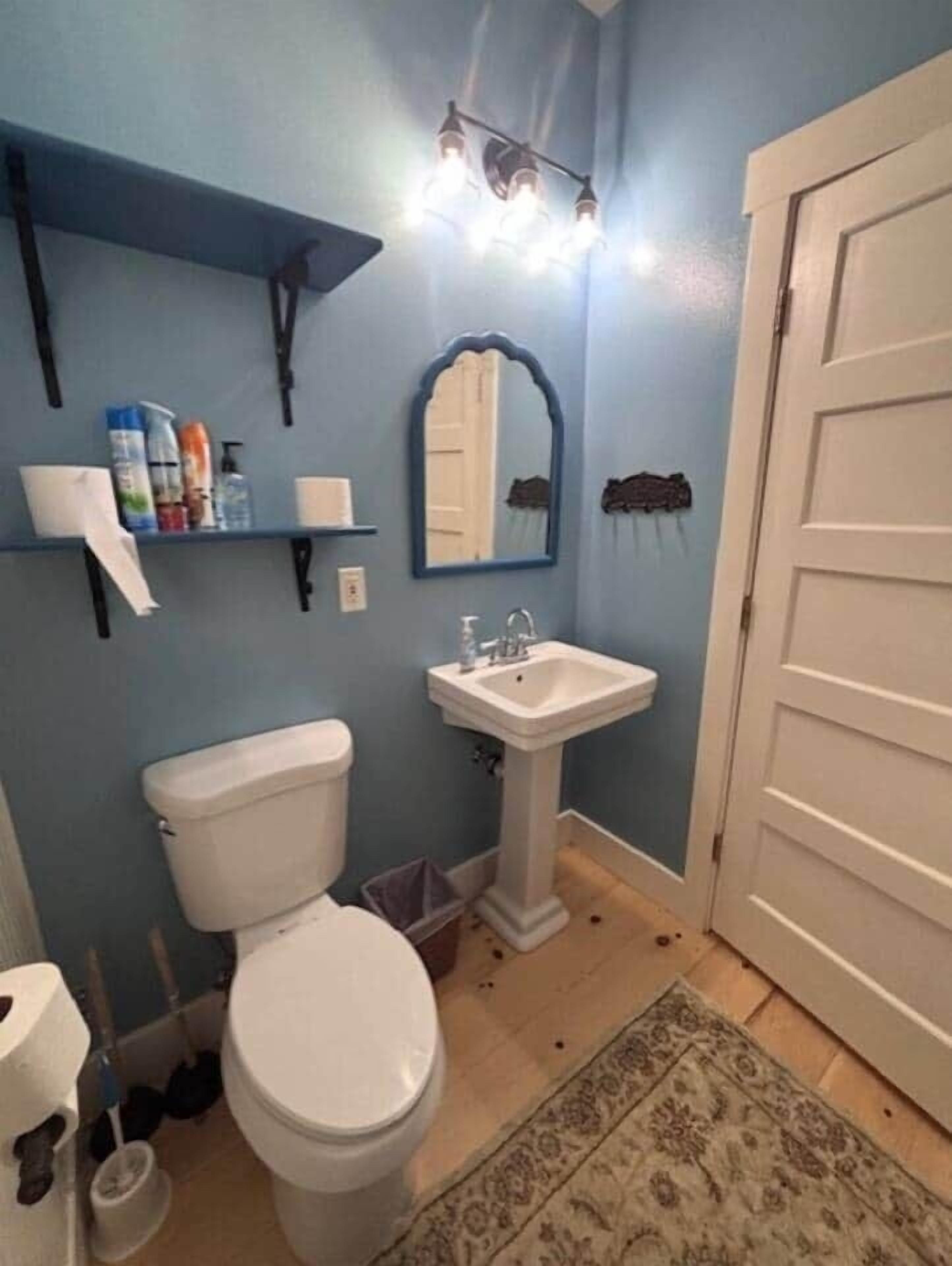 Baño con inodoro, lavamanos, espejo y repisas con artículos de aseo.