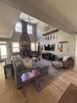 A spacious living room with a stone fireplace, a large sofa, a television, and a ceiling fan. - Yosemite Firehouse Inn-Cottage (Mariposa County)