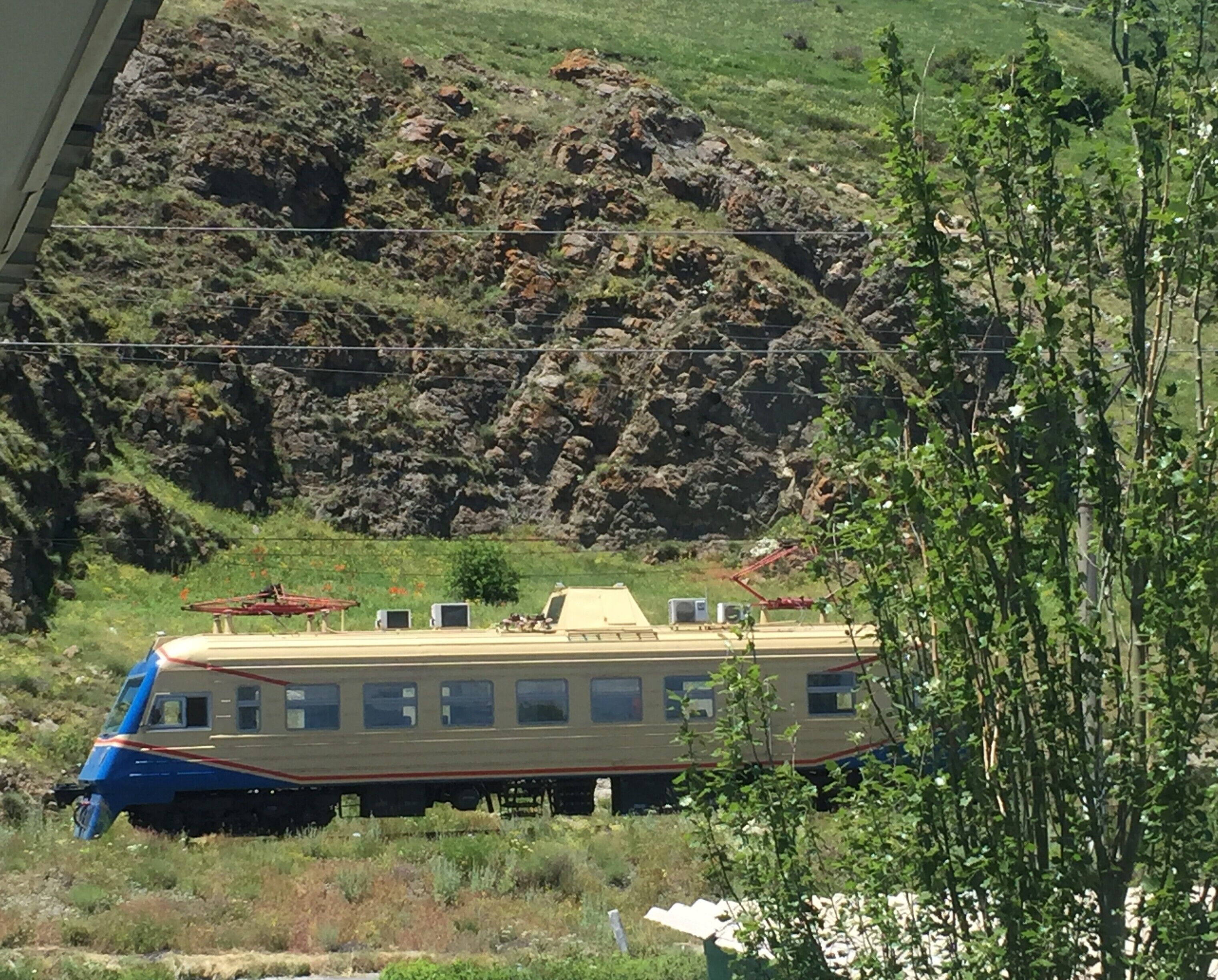 Un tren con múltiples ventanales detenido sobre una vía, rodeado de terreno rocoso y vegetación.