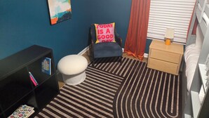 A room with a striped rug, a chair with a pillow, a bedside table with a lamp, a bookshelf, and a window with blinds. - 5 Min to Downtown • Hot Tub • Game Room (Atlanta)