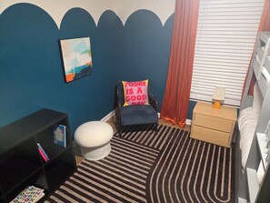 A room with a striped rug, a chair with a pillow, a bedside table with a lamp, a bookshelf, and a window with blinds. - 5 Min to Downtown • Hot Tub • Game Room (Atlanta)