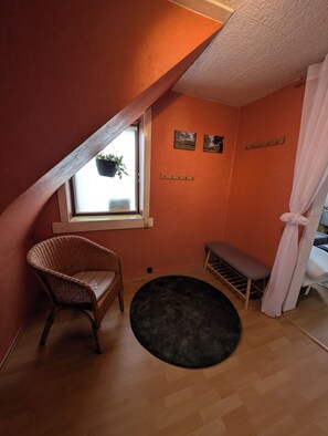 A room with a sloped ceiling, a wicker chair, a bench, a black rug, and a window with a hanging plant. - Gemütliche & familienfreundliche Ferienwohnung von HerzZeit- Harz in Wernigerode (Wernigerode)