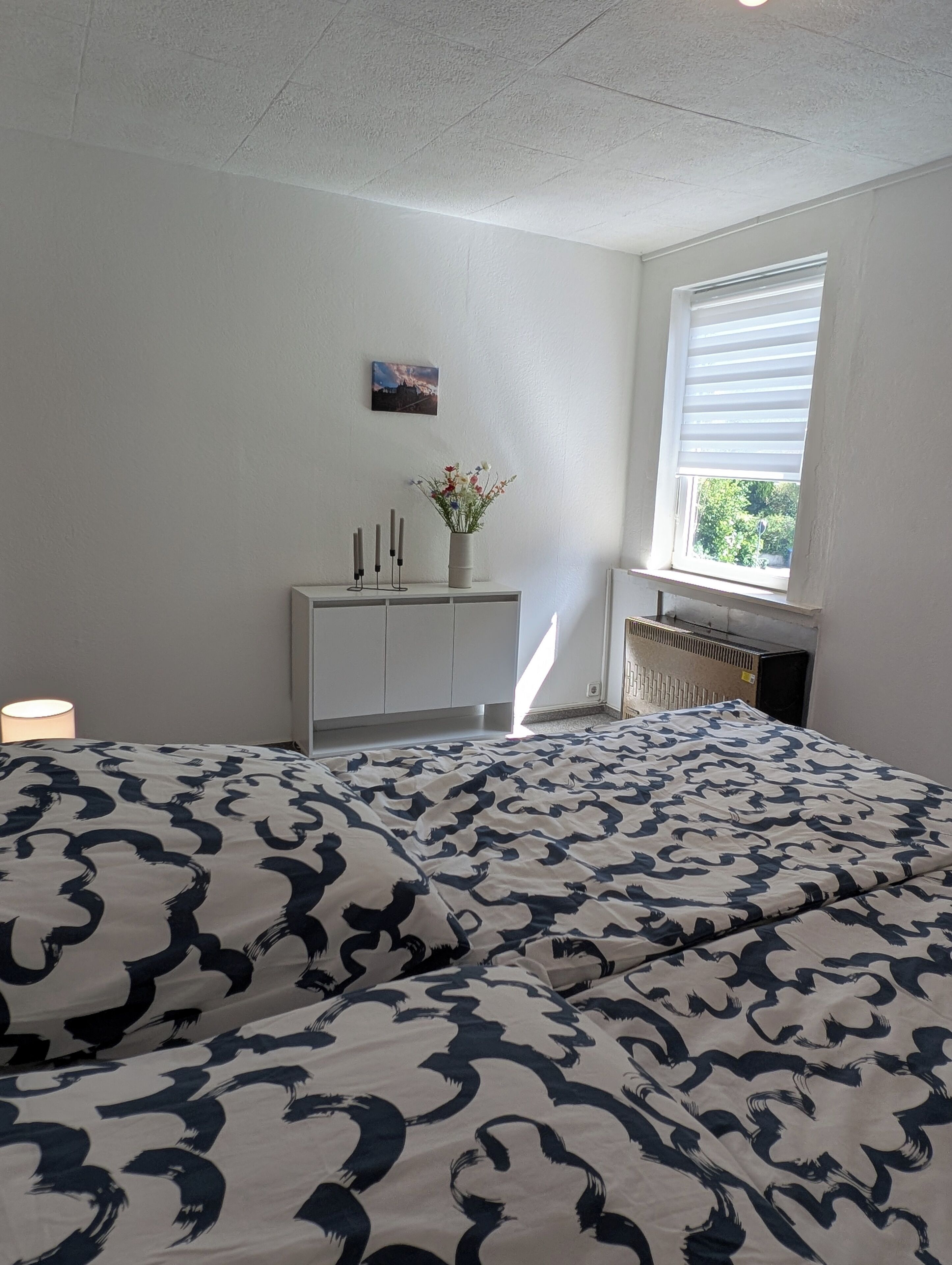 A bedroom with a bed, a white dresser, a window with blinds, and a radiator.