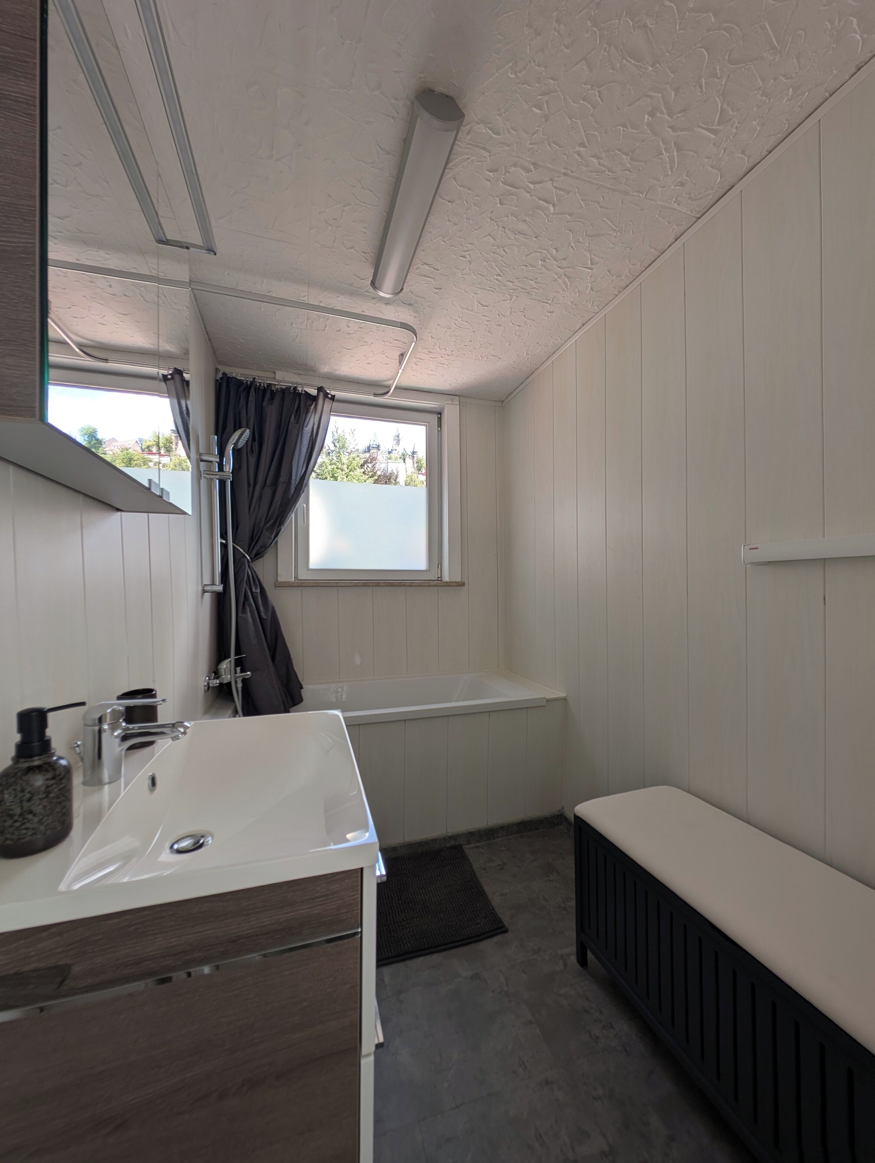 A bathroom with a white sink, a bathtub, a window with curtains, and a bench.