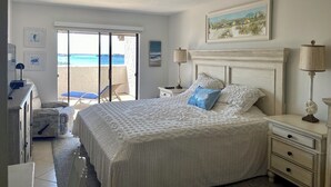 Ein Schlafzimmer mit einem großen Bett, einem Schreibtisch, einem Sessel und Blick auf den Strand durch eine Glastür.