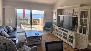 Ein Wohnzimmer mit Sofa, Sesseln, einem Couchtisch, einem Fernseher und einem Schrank. Eine Glastür führt auf eine Außenpatio mit Tisch und Stühlen.