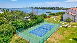 Ein Tennisplatz, umgeben von Grünflächen, mit Blick auf ein Gewässer und Gebäude in der Ferne.