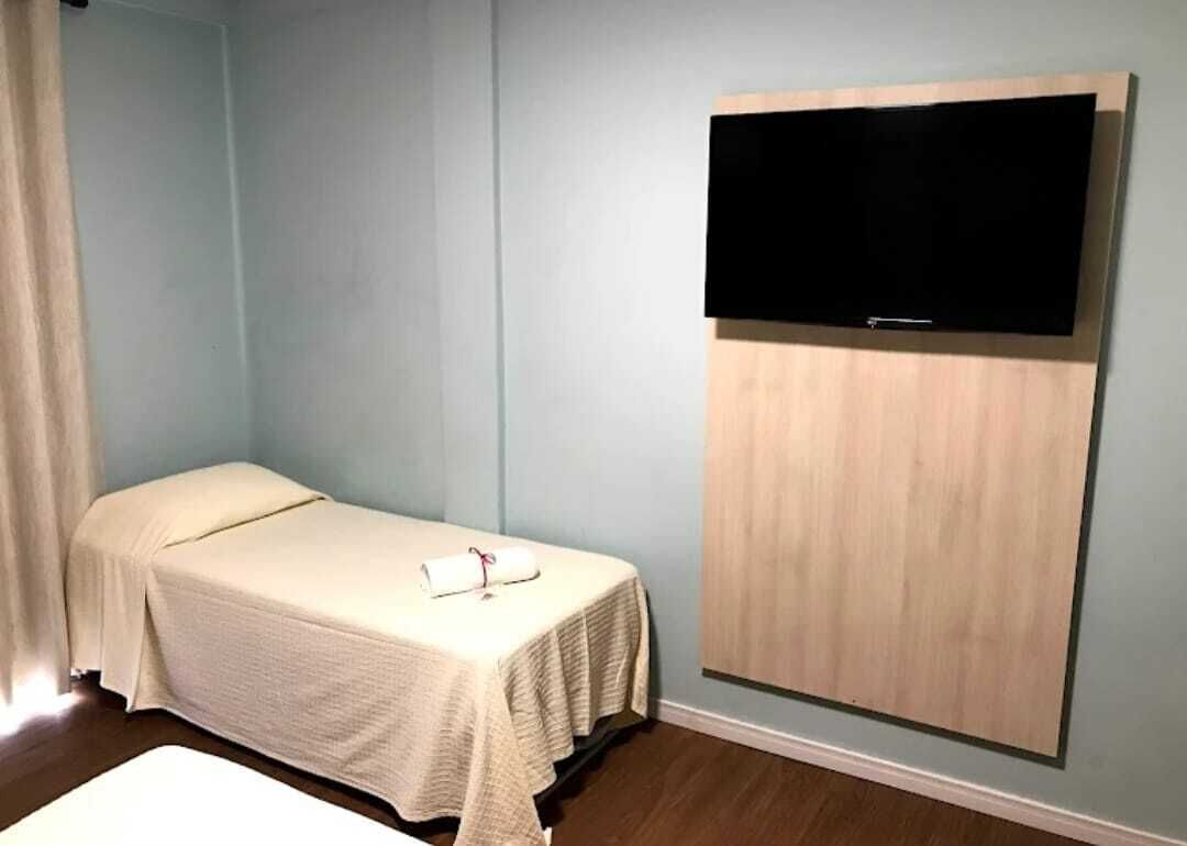 Un letto singolo con coperta bianca, una TV a schermo piatto montata su un pannello di legno e una parete azzurra.