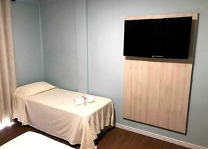 Una cama individual con cubierta blanca, un televisor de pantalla plana montado sobre un panel de madera y una pared de color azul claro.