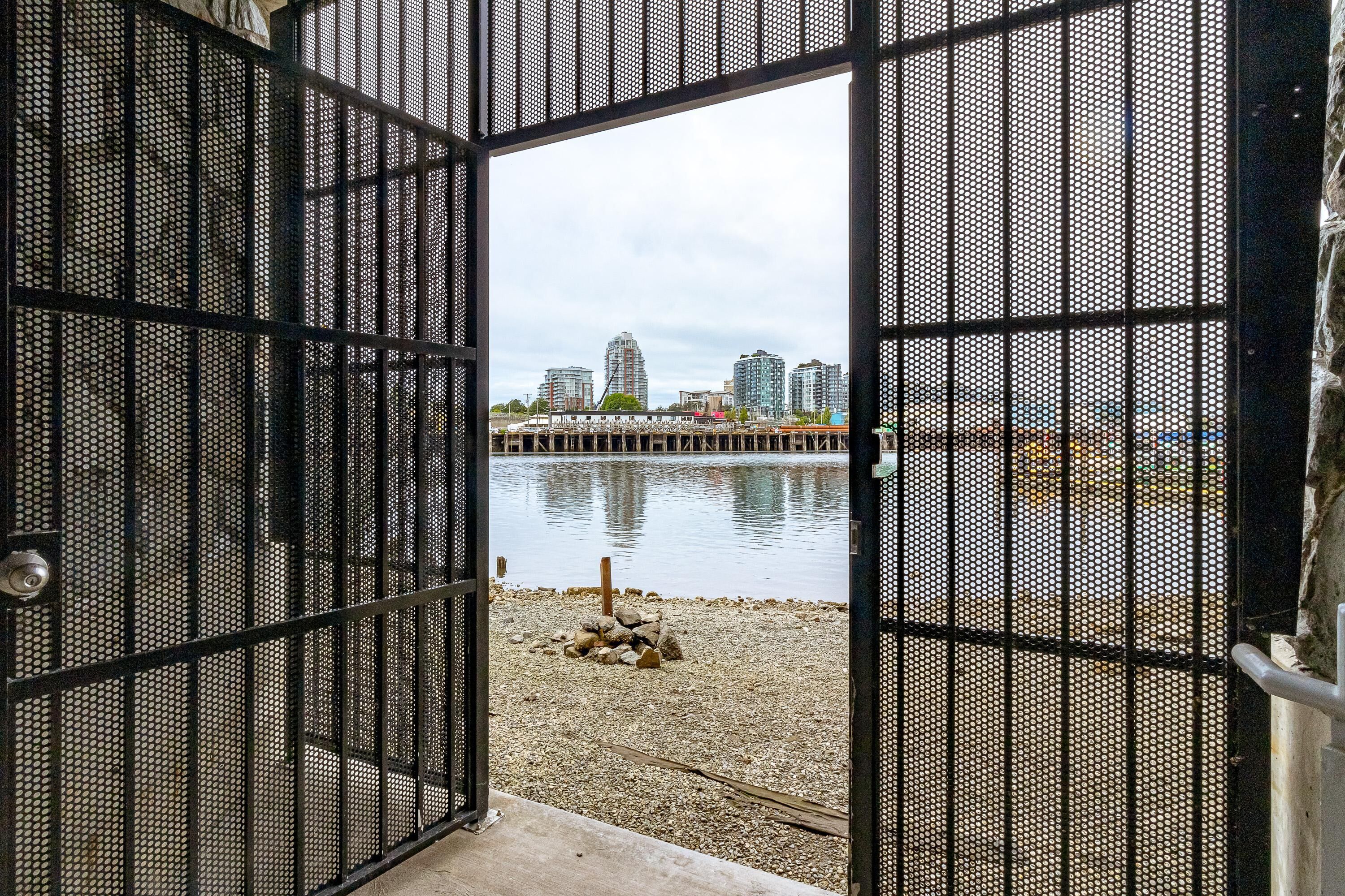 An open metal gate leading to a pebbled waterfront with buildings in the background.
