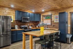 A modern kitchen with wooden cabinets, stainless steel appliances, a wooden dining table, and bar stools. - Renovated Playhouse—Slide/Views/Fire Pit/Hot Tub (Pigeon Forge)