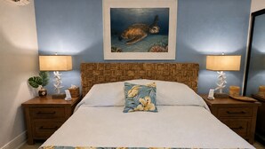 A bedroom with a bed, nightstands, a painting of a sea turtle, and a floral bedspread. - Oceanfront, swim with turtles, enjoy sunset views, all from your backyard. #B18 (Lahaina)