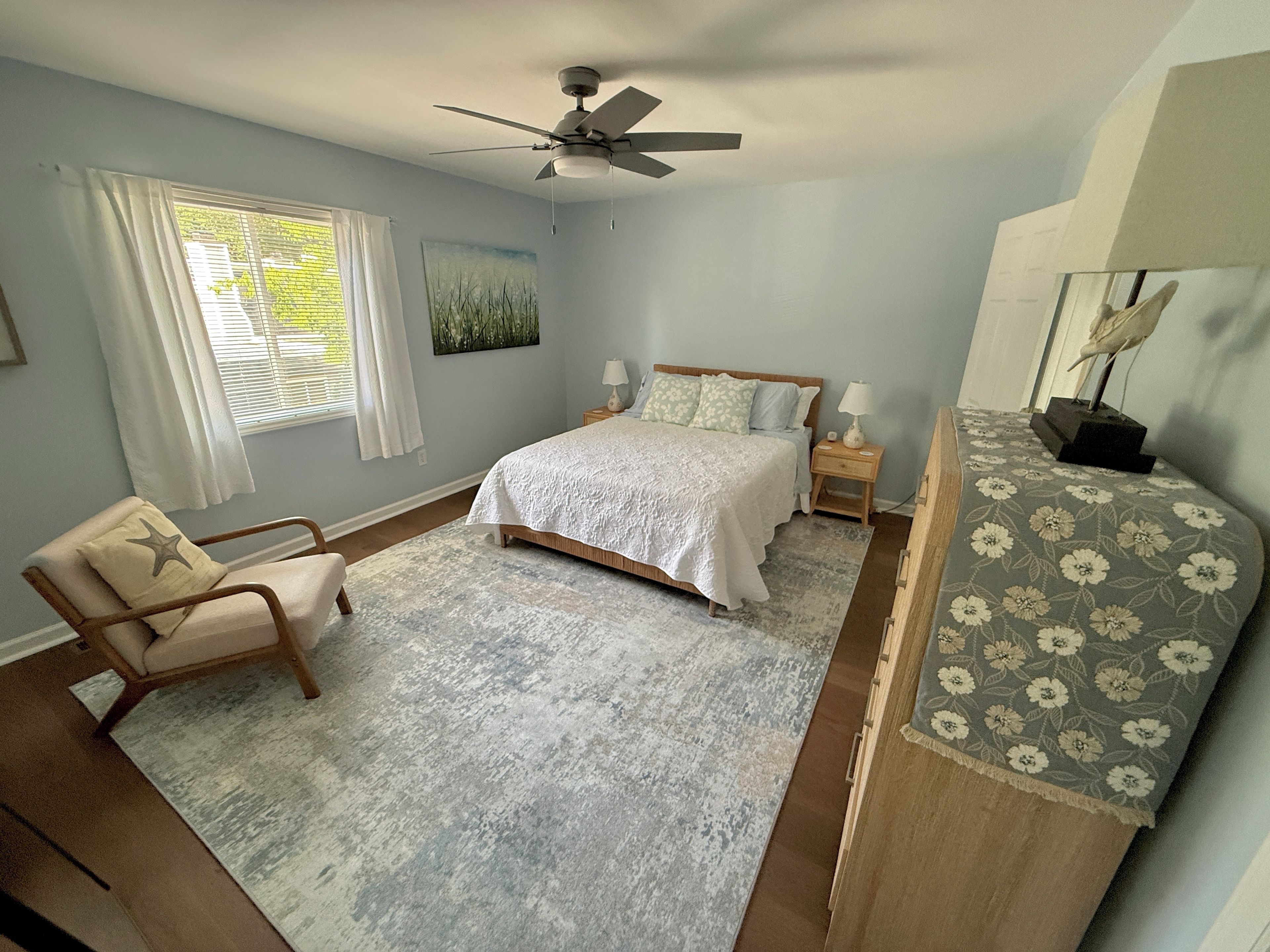 A bedroom with a bed, a chair, a nightstand, a ceiling fan, and a window with curtains.