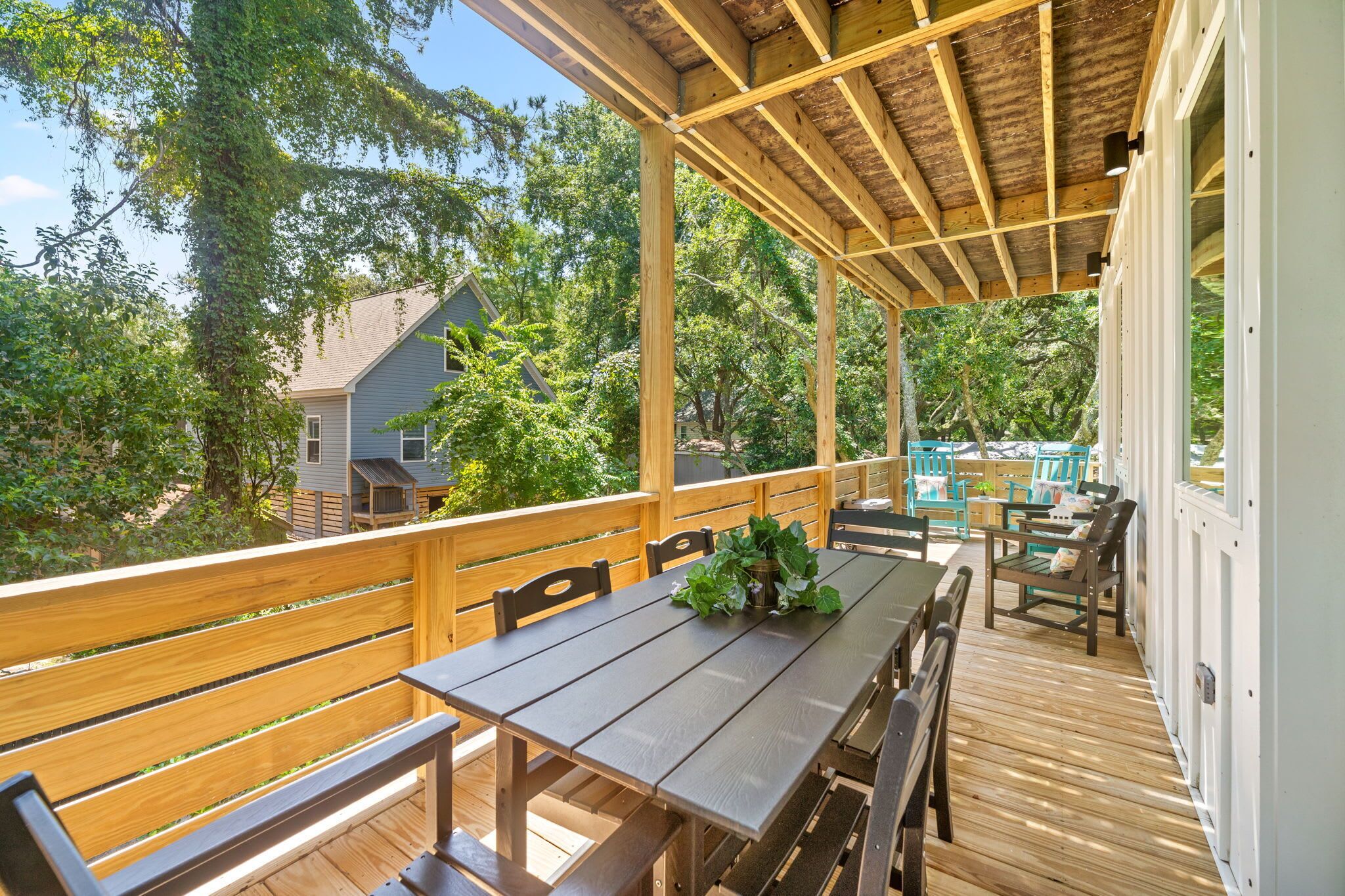 A covered outdoor deck with a table and chairs, surrounded by wooden railings, and overlooking a residential area with trees.