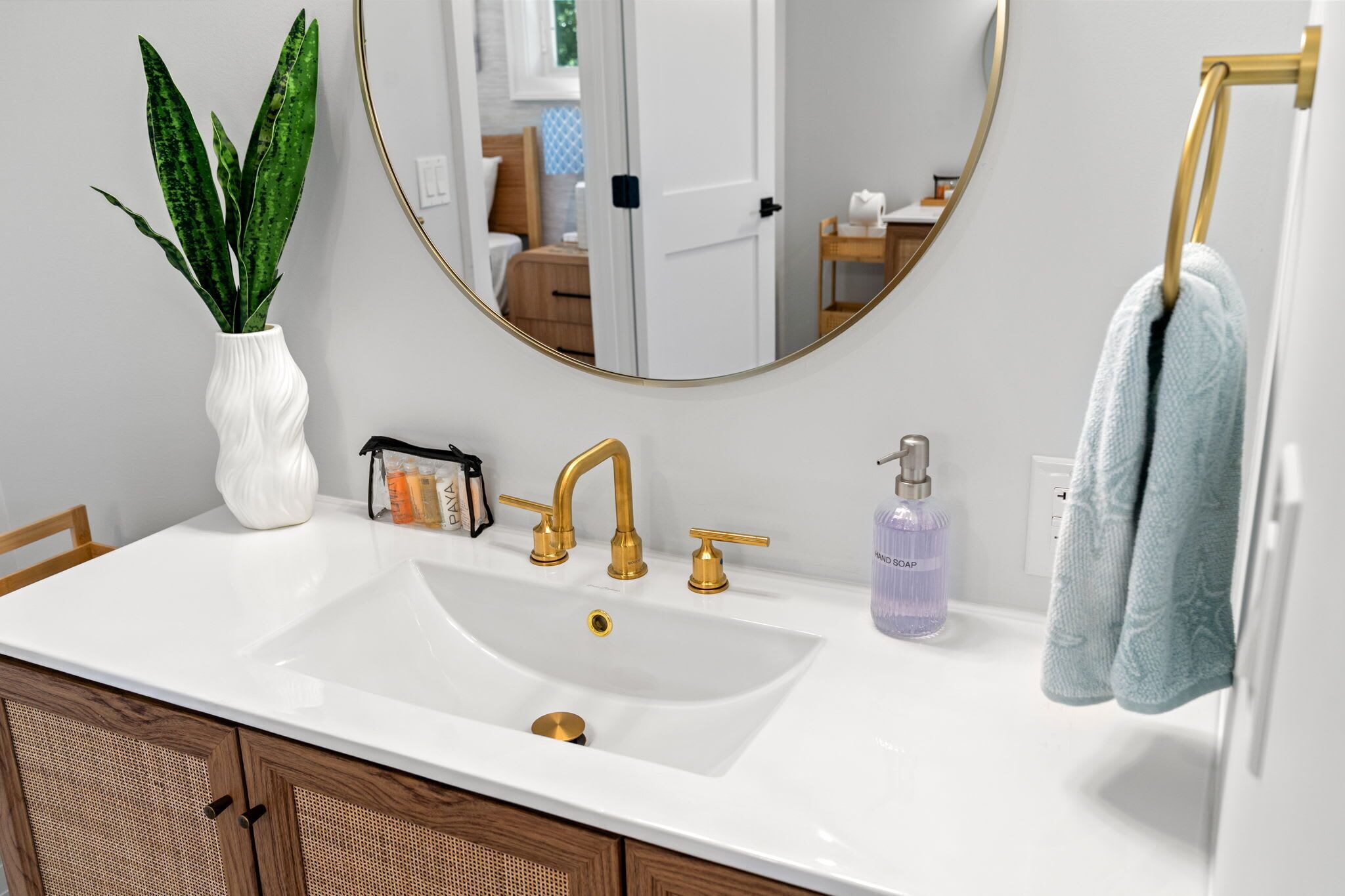 A bathroom with a white sink, gold faucets, a round mirror, a potted plant, a towel rack, and a soap dispenser.