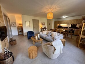 Ein großzügiger Wohnbereich mit einer Couch, Sesseln, einem Couchtisch und einem Esstisch. Im Hintergrund befindet sich eine Küche mit Holzschränken und einer Pendelleuchte.