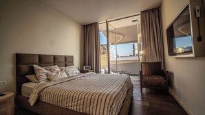 Une chambre d’hôtel avec un grand lit, une table de chevet, une chaise, une télévision et un balcon avec des rideaux.