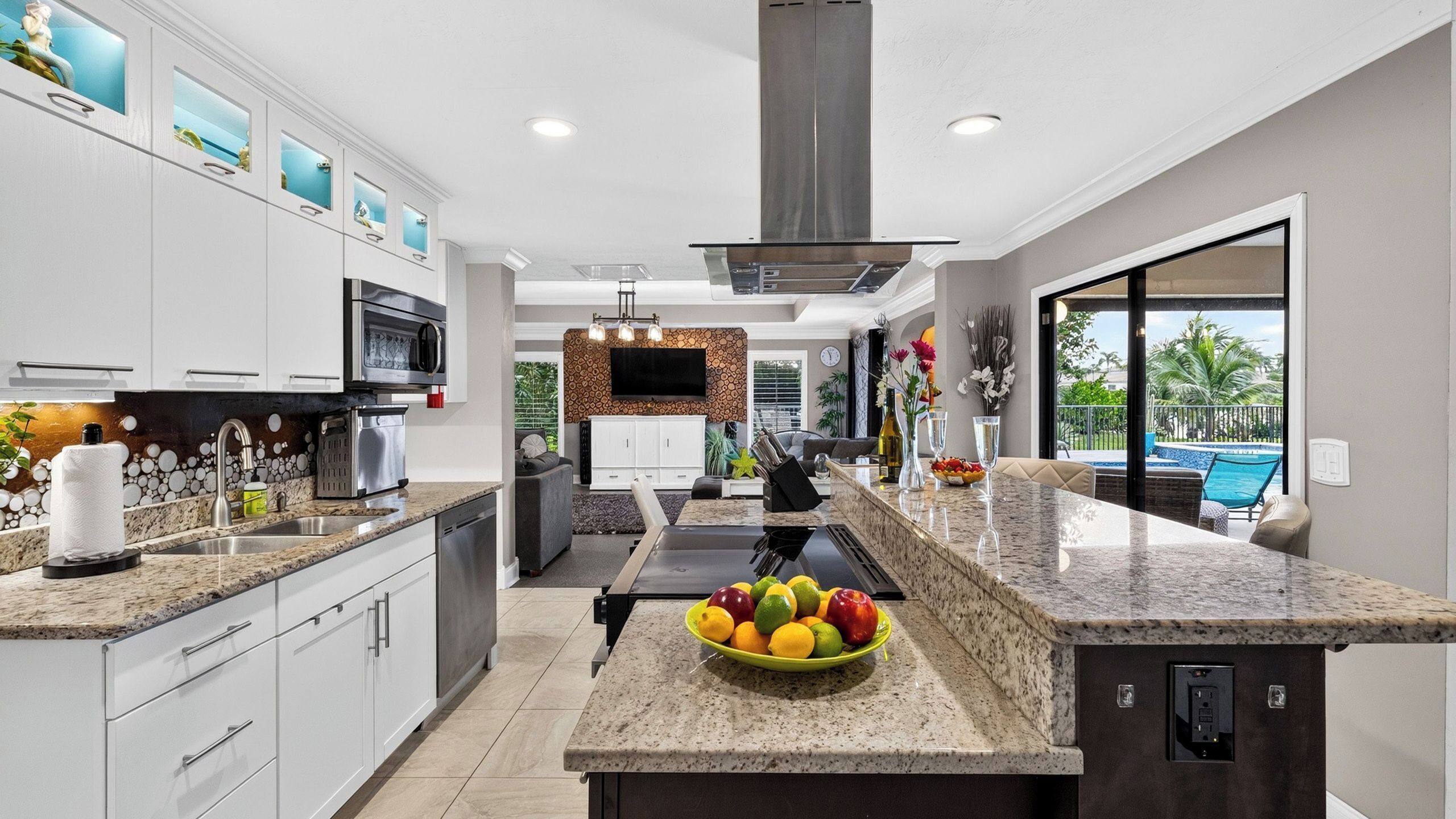 A modern kitchen with granite countertops, a central island with fruit, and a view of a swimming pool.