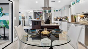A modern kitchen with a glass dining table, white chairs, stainless steel appliances, and decorative elements. - Blue Sunset by Avantstay Pool + Spa + Fire Table Near Beaches + Naples (Bonita Springs)