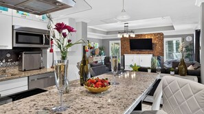A modern kitchen with a granite countertop, stainless steel appliances, and a dining area with a white upholstered chair. - Blue Sunset by Avantstay Pool + Spa + Fire Table Near Beaches + Naples (Bonita Springs)