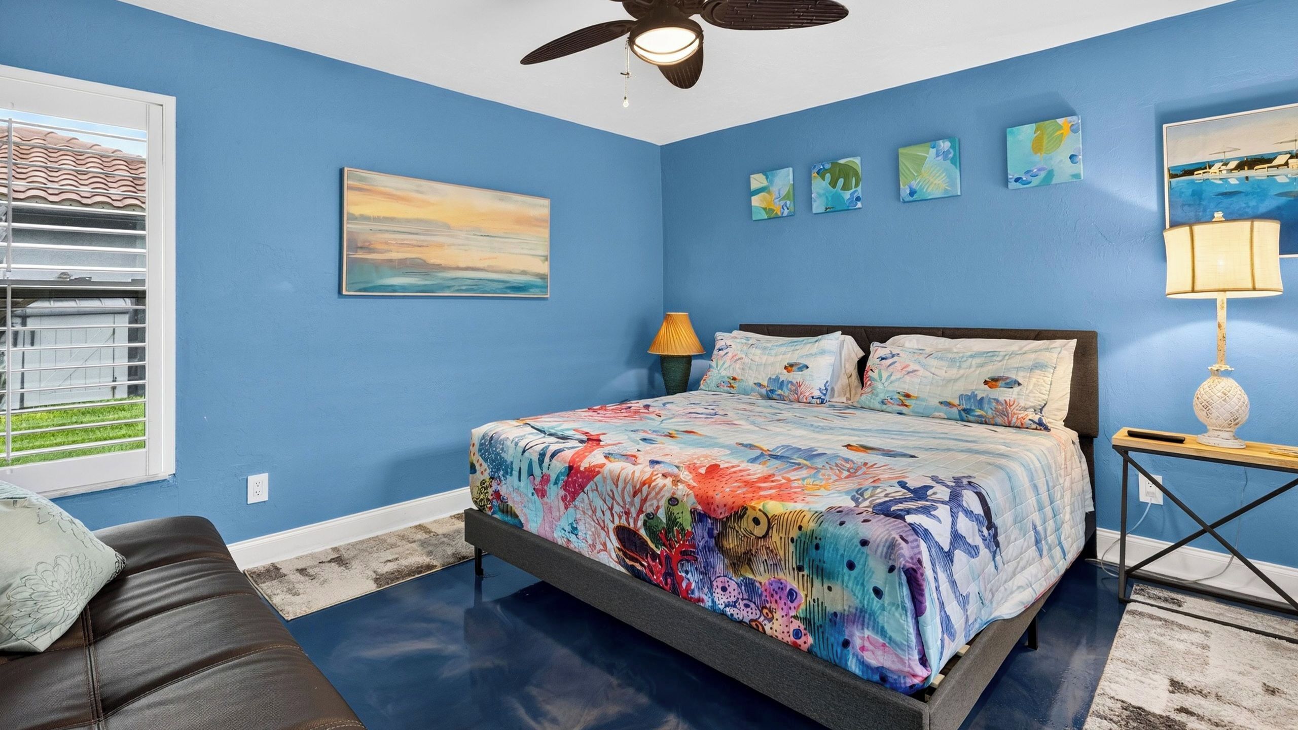 A bedroom with a bed, a ceiling fan, a window with blinds, and artwork on the walls.