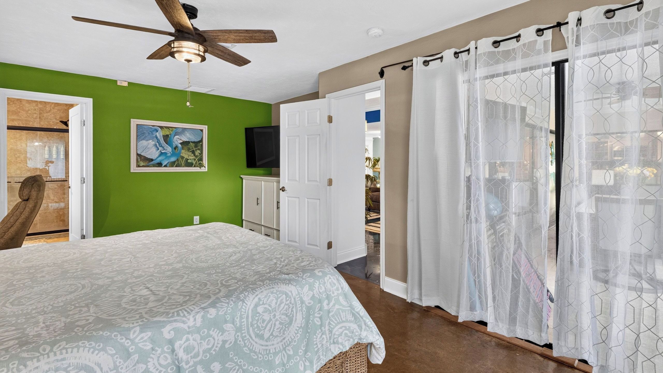 A bedroom with a bed, a ceiling fan, a green wall, a painting, a television, and a door leading to another room.