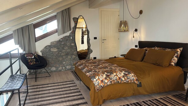 Quarto com uma cama grande, parede de pedra, espelho, cadeira e vista para o exterior.