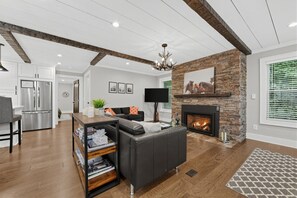 A modern living room with a fireplace, a black leather sofa, a wooden coffee table, and a kitchen area with a stainless steel refrigerator.