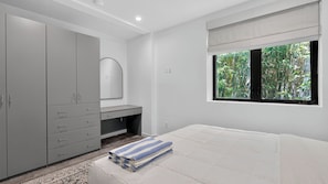 A bedroom with a bed, a grey wardrobe, a vanity with a mirror, and a window with a view of greenery. - Canal Crown by Avantstay Rooftop Lounge + Grill Venice Canals + Abbot Kinney (Venice)