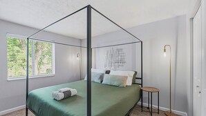 A four-poster bed with a green bedspread, a round side table, a floor lamp, and a window with blinds. - King Canopy Bed I Sofa Bed I Next to Restaurants (Kettering)