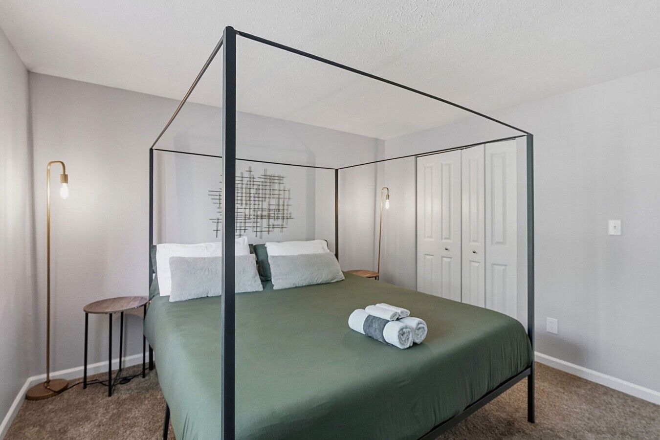 Una cama con dosel con una colcha verde y almohadas blancas, una mesita redonda con una lámpara, y una lámpara de pared.