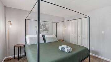 Una cama con dosel con una colcha verde y almohadas blancas, una mesita redonda con una lámpara, y una lámpara de pared.