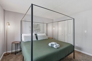 Un lit à baldaquin avec une couvre-lit verte et des oreillers blancs, une petite table ronde avec une lampe et un luminaire fixé au mur.