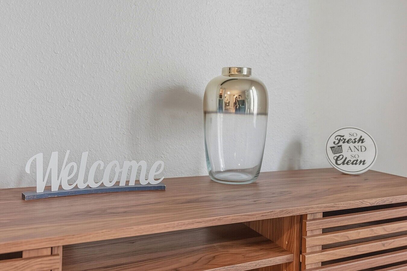A wooden table with a clear glass vase and a 'Welcome' sign.