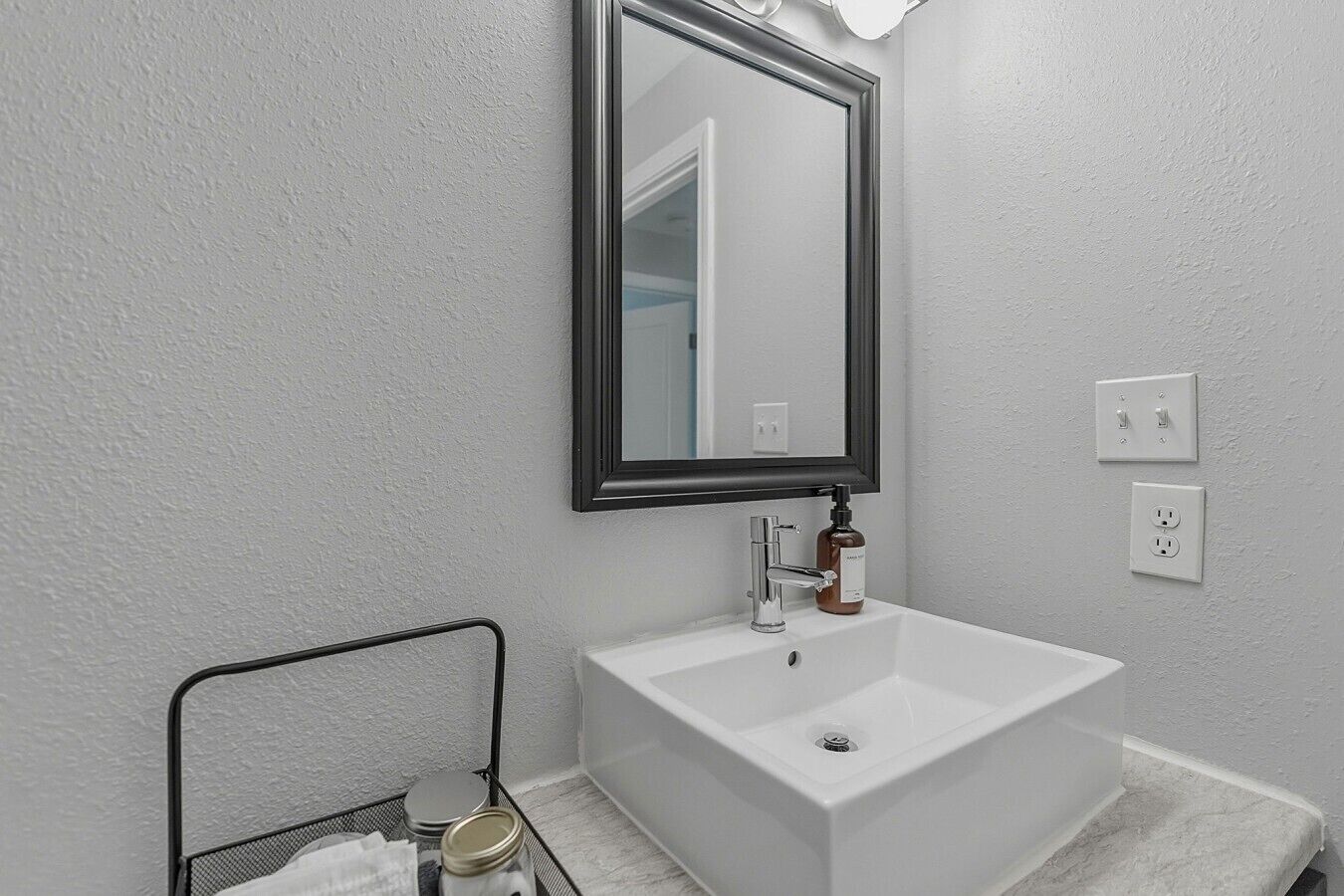 A bathroom with a white sink, a mirror, and a metal rack with jars.