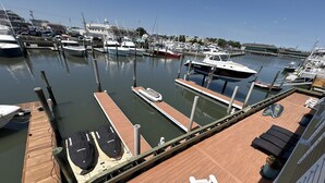 Un puerto deportivo con varias embarcaciones amarradas, una terraza de madera con sillones de descanso, y vista al puerto en el fondo.