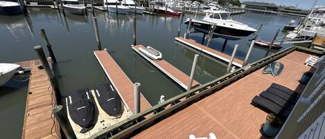 Un puerto deportivo con varias embarcaciones amarradas, una terraza de madera con sillones de descanso, y vista al puerto en el fondo.