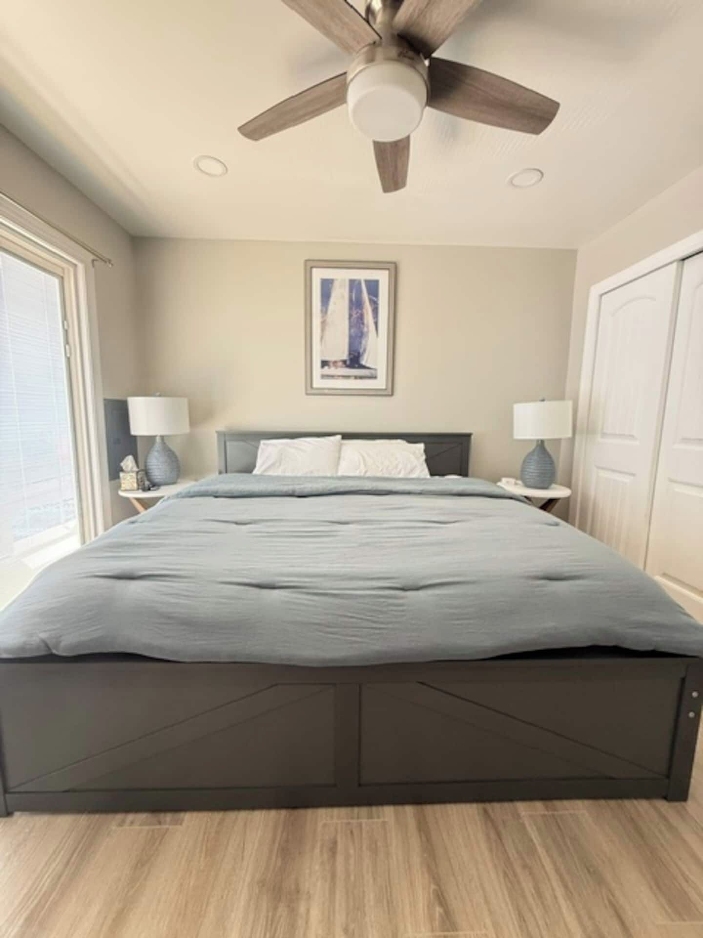 Une chambre à coucher avec un lit, deux tables de chevet, un ventilateur de plafond, un tableau encadré au mur et une fenêtre.